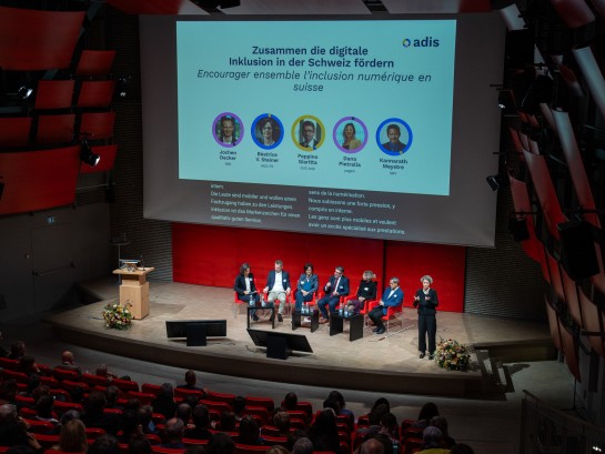 Alle Podiumsdiskussionsteilnehmer sitzen auf der Bühne. Hinter ihnen sind sie auf der Leinwand mit Foto zu sehen. Von links nach rechts sitzen: Jochen Decker, Béatrice Vatron-Steiner, Peppino Giaritta, Dana Pietralla und Kannarath Meystre.