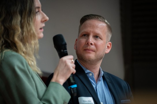 Sandrine Denti von der digitalswitzerland foundation redet in ein Mikrofon. Dahinter hört ihr Christian Maag vom Dachverband Lesen und Schreiben gebannt zu.