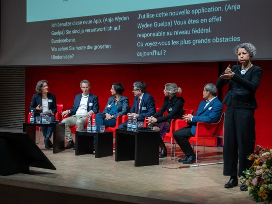 Alle Podiumsdiskussionsteilnehmer sitzen auf der Bühne. Von links nach rechts sitzen: Jochen Decker, Béatrice Vatron-Steiner, Peppino Giaritta, Dana Pietralla und Kannarath Meystre. Im Vordergrund ist die Übersetzerin für Gebärdensprache zu sehen.