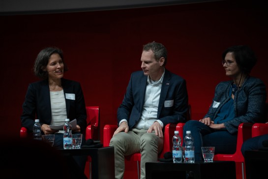 Anja Wyden Guelpa pose une question aux participants de la table ronde, Jochen Decker et Béatrice Vatron-Steiner. Les trois participants sont assis sur scène sur des chaises rouges. Devant eux, des bouteilles d'eau sont posées sur des tables basses noires