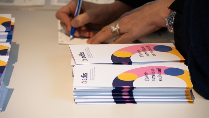 Une main écrivant sur du papier avec un stylo à bille bleu. À côté, des piles de brochures ADIS avec l’inscription L’avenir numérique est inclusif et un design coloré composé d’éléments circulaires jaunes, roses, bleus et blancs.